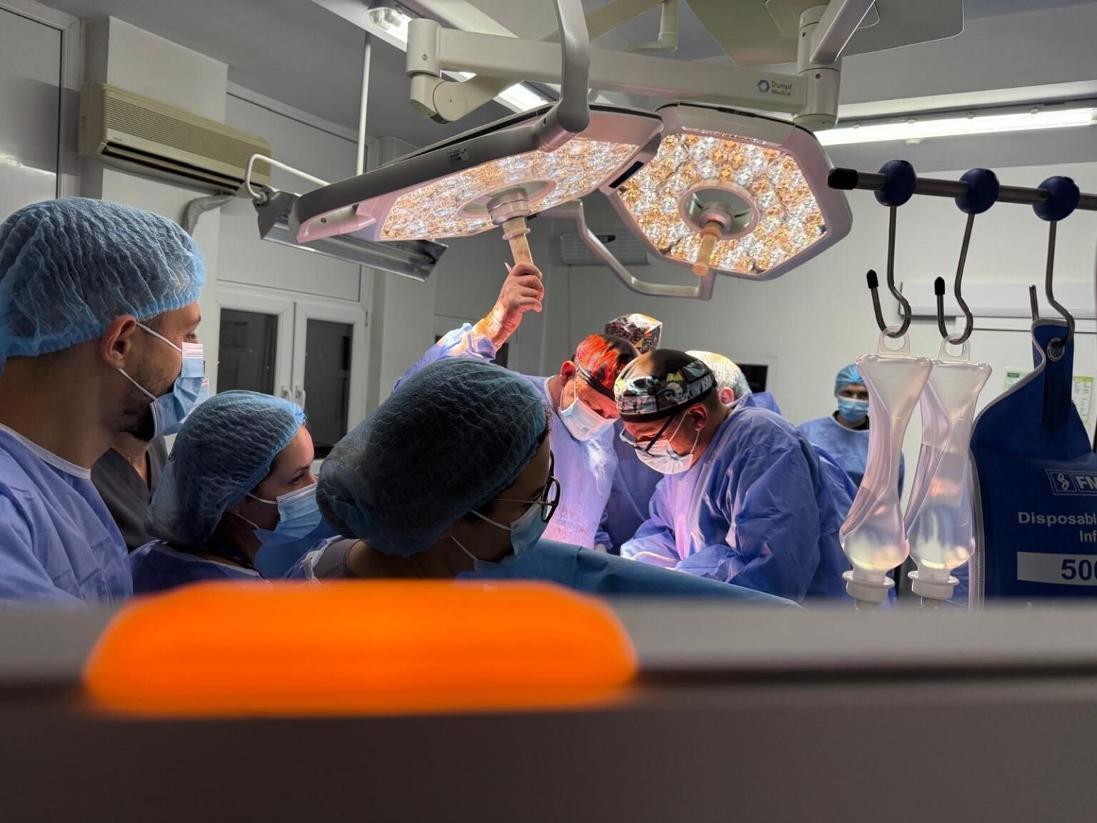 Хирурги в синих халатах и масках оперируют под ярким светом хирургических ламп