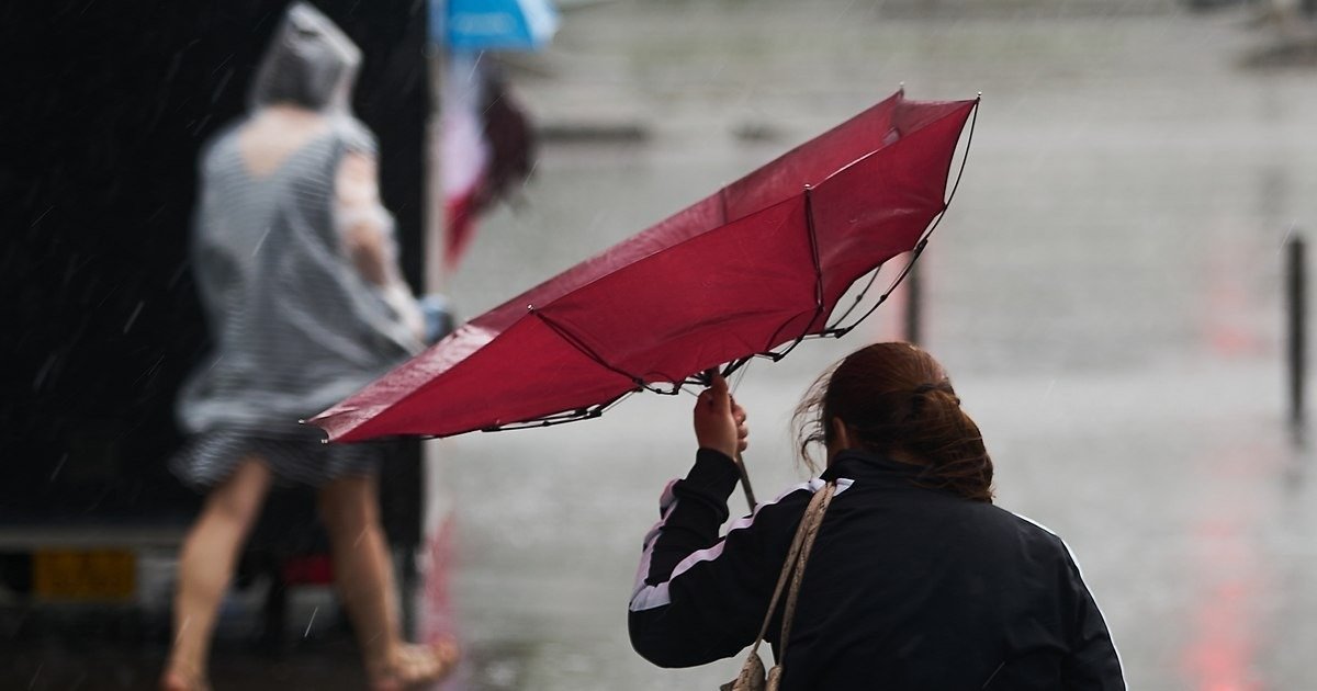Женщина пытается удержать вывернутый ветром красный зонт под проливным дождём на городской улице
