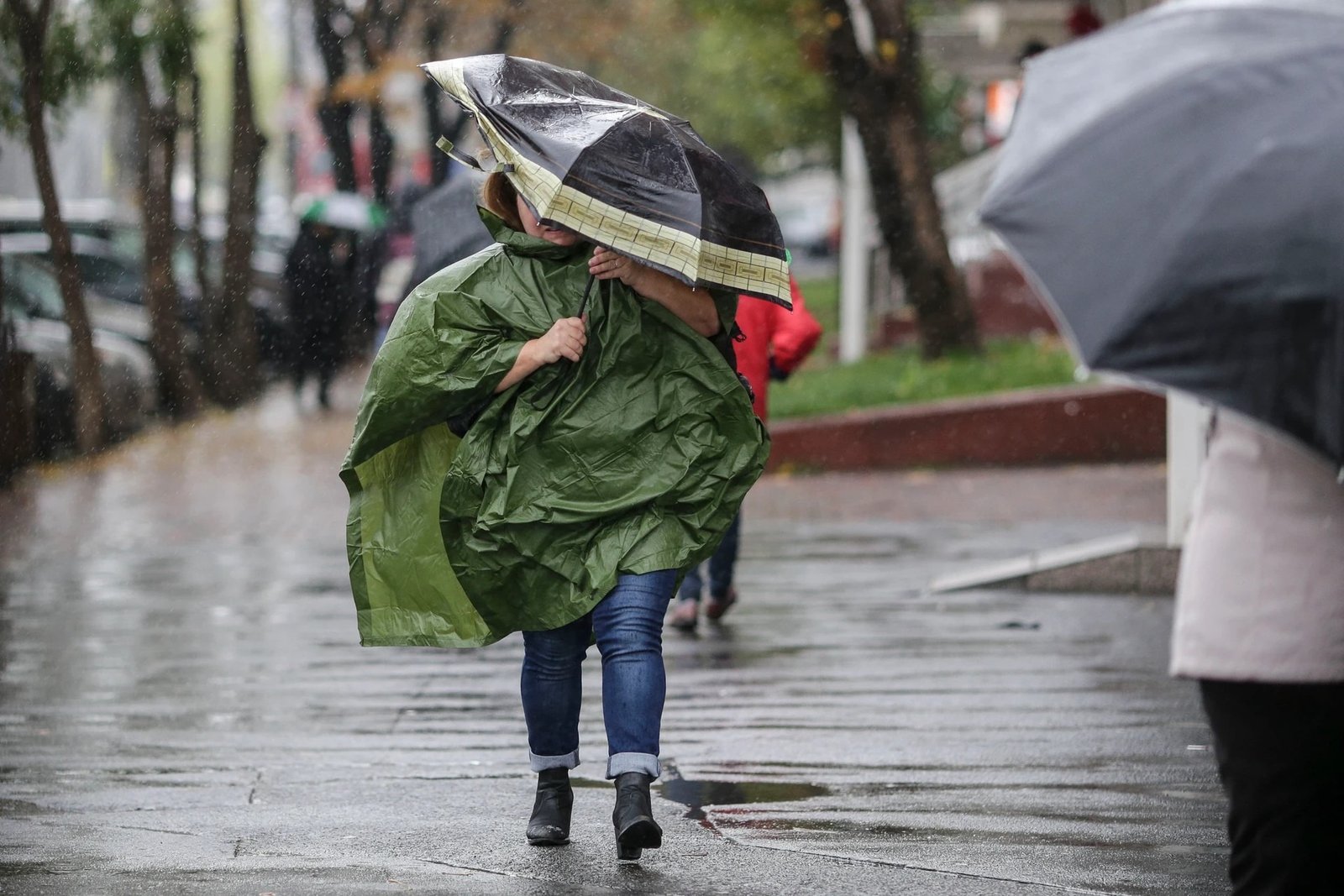 Пешеход в зелёном дождевике идёт по мокрой улице под сильным ливнем, укрываясь зонтом
