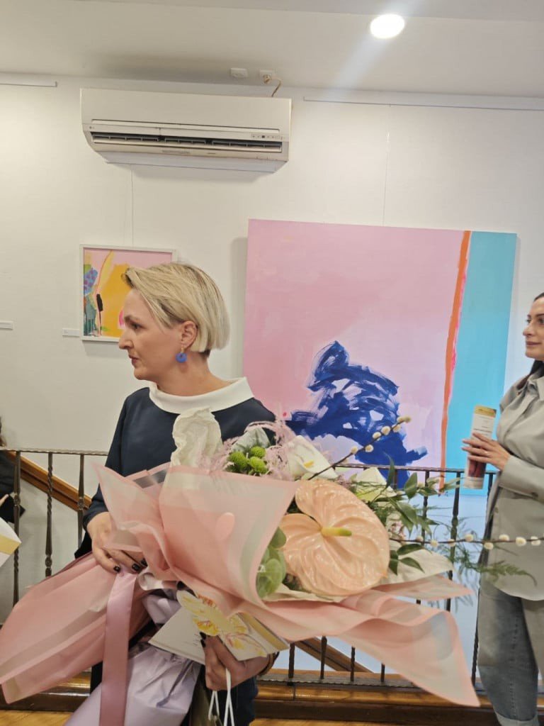 Художница Ивона Скочилич с букетом цветов на фоне одной из своих работ розово-голубой палитры — на открытии персональной выставки в Мостаре