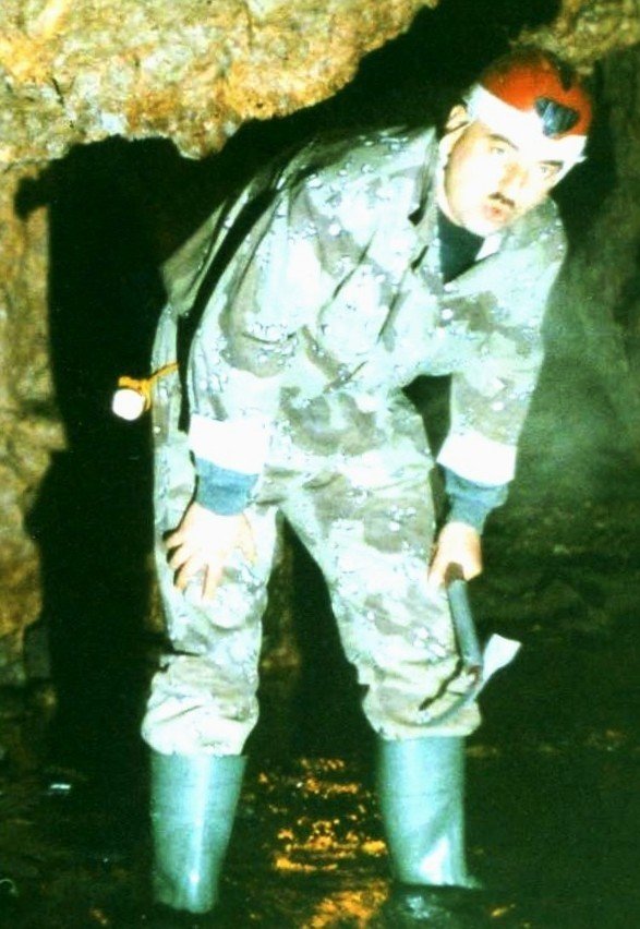 Спелеолог в комбинезоне и каске с налобным фонарём осматривает свод карстовой полости в горах Горжа, держа в руке геологический молоток