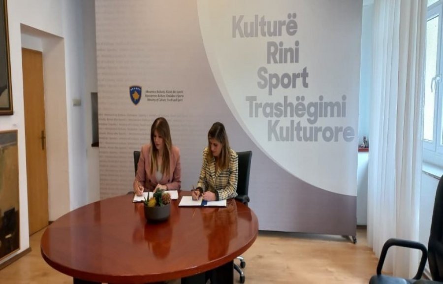 Две женщины подписывают документы за круглым деревянным столом в зале Министерства культуры Косово под гербом республики и надписью «Kulturë Rini Sport Trashëgimi Kulturore»
