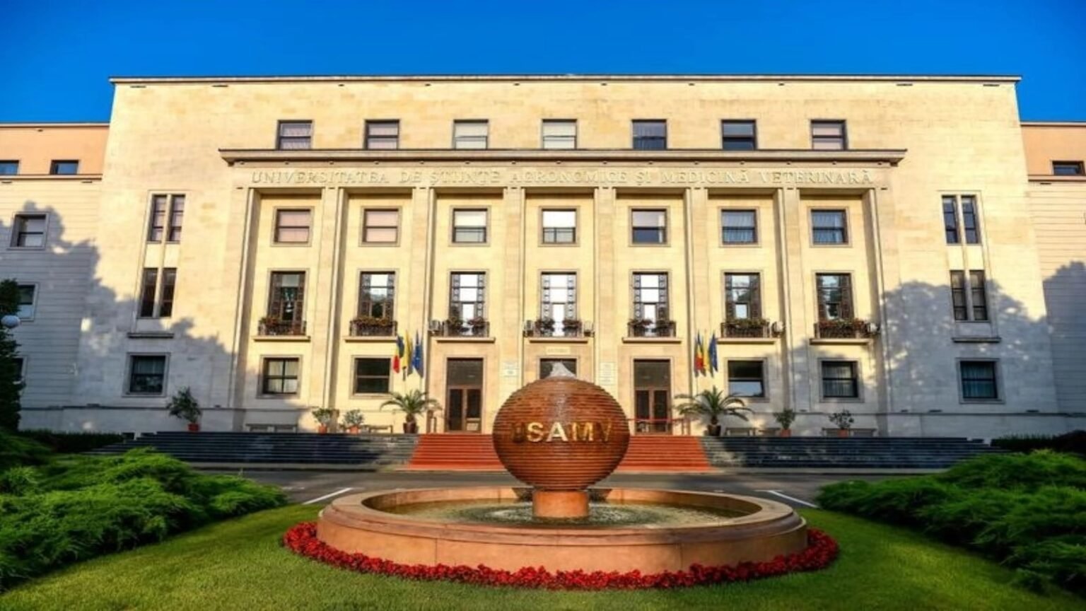 Главный исторический корпус Университета аграрных наук и ветеринарной медицины (USAMV) в Бухаресте: каменный фасад с надписью «UNIVERSITATEA DE ȘTIINȚE AGRONOMICE ȘI MEDICINĂ VETERINARĂ», два румынских флага у входа и фирменный фонтан-глобус «USAMV» с клумбой красных цветов. Здесь открылась XV международная конференция Agriculture for Life — Life for Agriculture