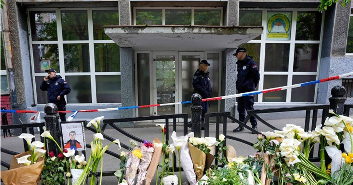 Сотрудники полиции у оцепленного входа в начальную школу «Владислав Рибникар» в Белграде, где в мае 2023 года несовершеннолетний сын Кецмановичей убил девятерых одноклассников и охранника; у решётки — фотография погибшей девочки и цветы