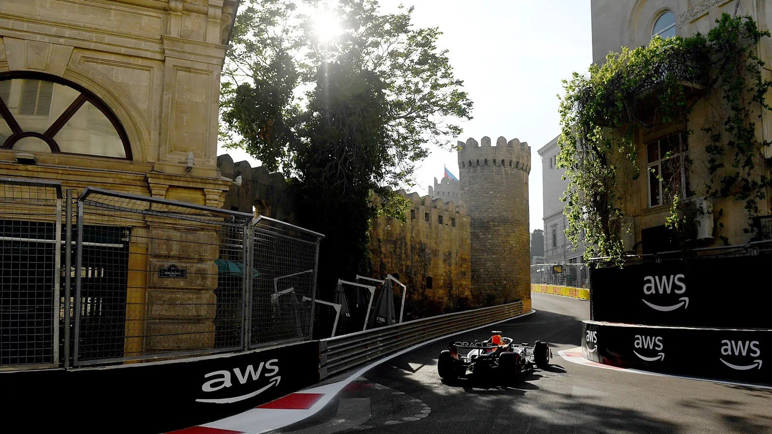 Болид Oracle Red Bull Racing выходит из поворота у стен старого города Ичери Шехер: слева — историческое здание, справа — крепостная стена с флагом Азербайджана и барьеры со спонсорской разметкой AWS