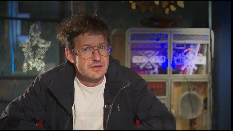 Крупный план кинооператора в очках и тёмной куртке во время интервью в помещении с приглушённым освещением и цветной подсветкой на фоне