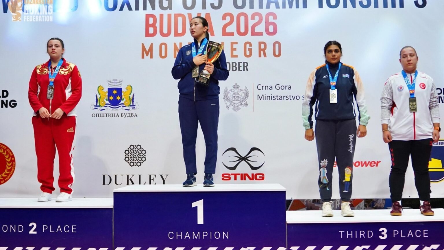 Церемония награждения на чемпионате мира по боксу U19 в Будве 2026: чемпионка на верхней ступени пьедестала, по бокам — серебряные и бронзовые призёры. На заднем плане видны логотипы Общины Будвы, Министерства спорта Черногории и спонсоров турнира